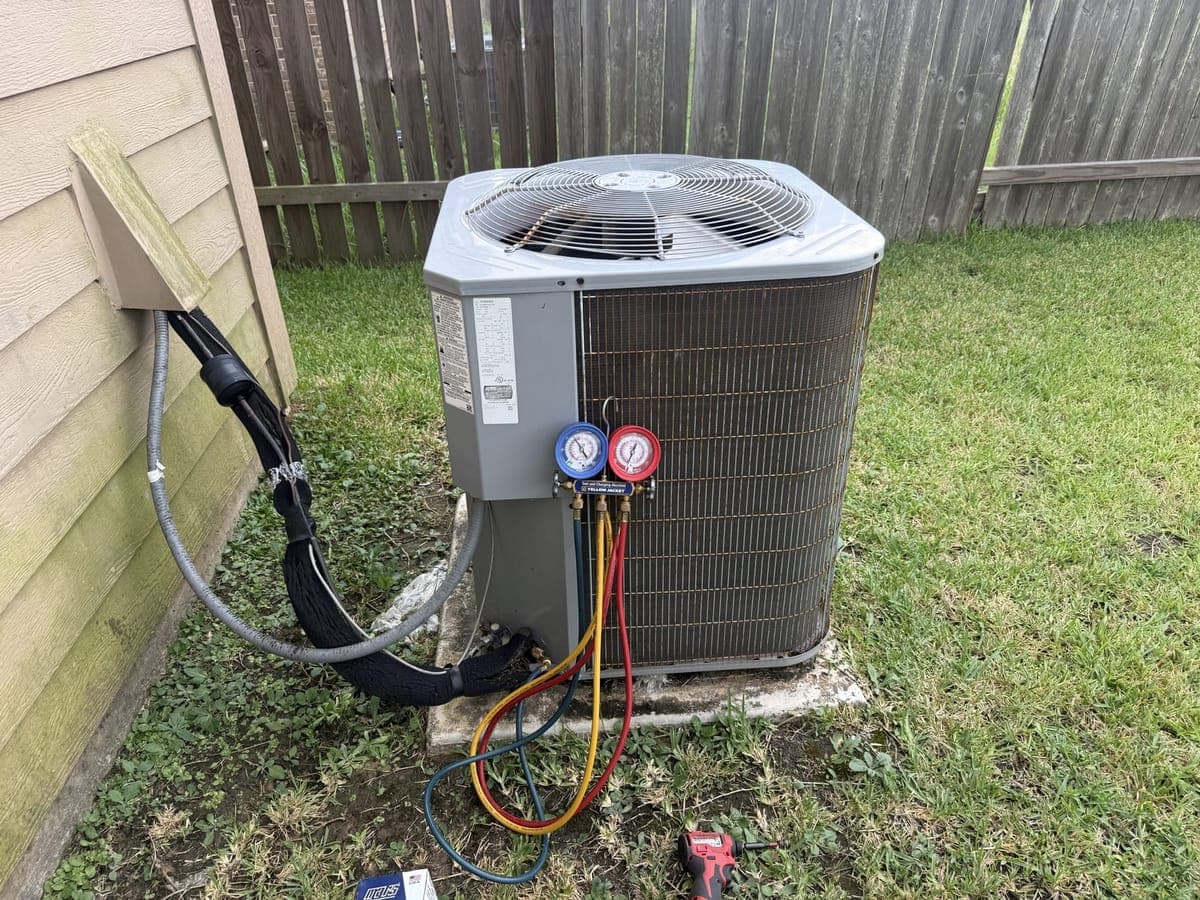 Outdoor AC unit beside a house with refrigerant gauges and hoses connected
