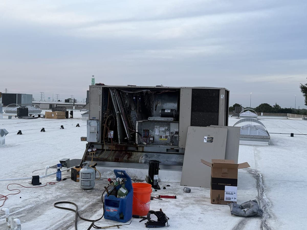 Commercial rooftop unit with its panels off on a flat white roof, with service equipment set out beside it