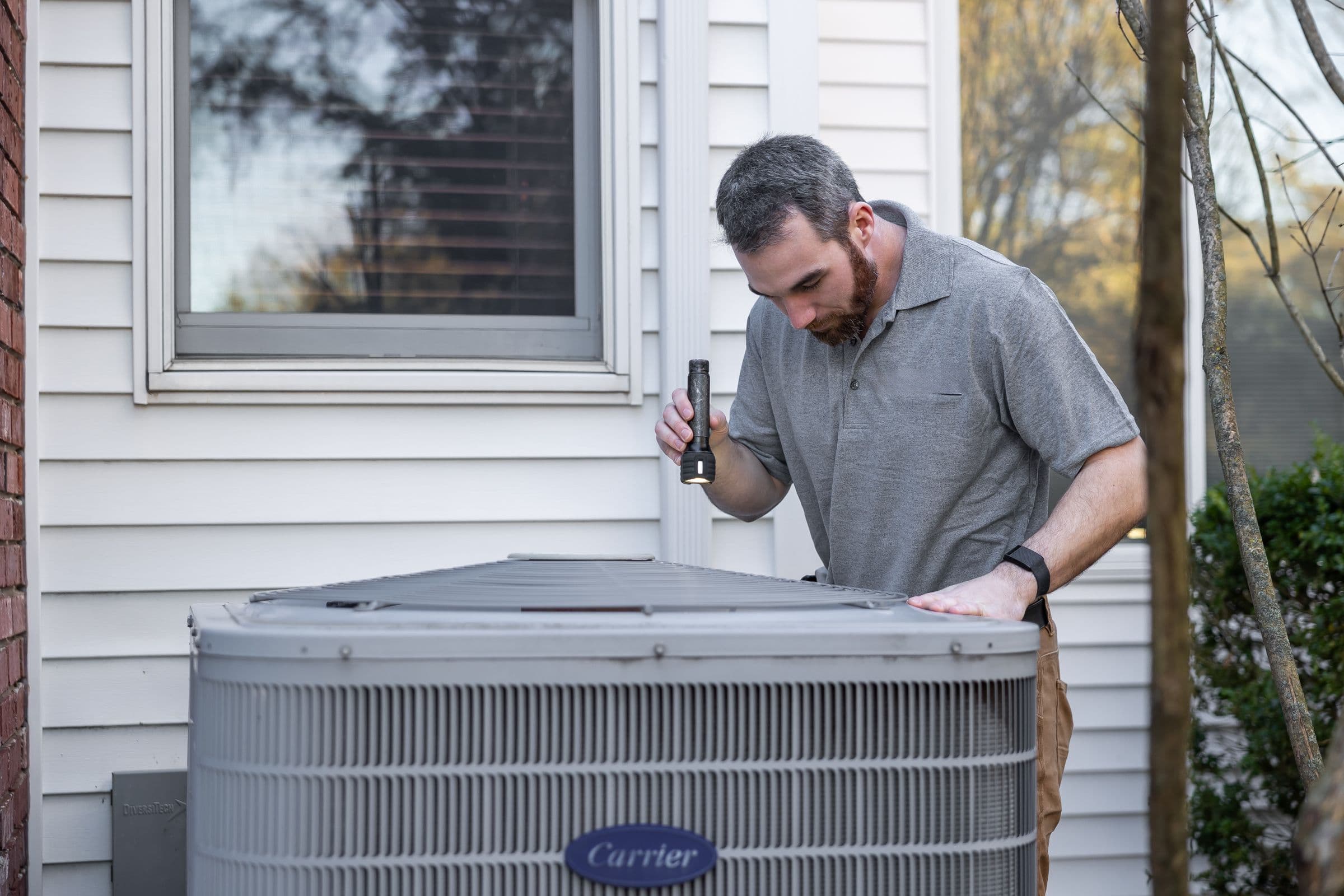 Technician shining a flashlight into a Carrier condenser beside the white siding of a house